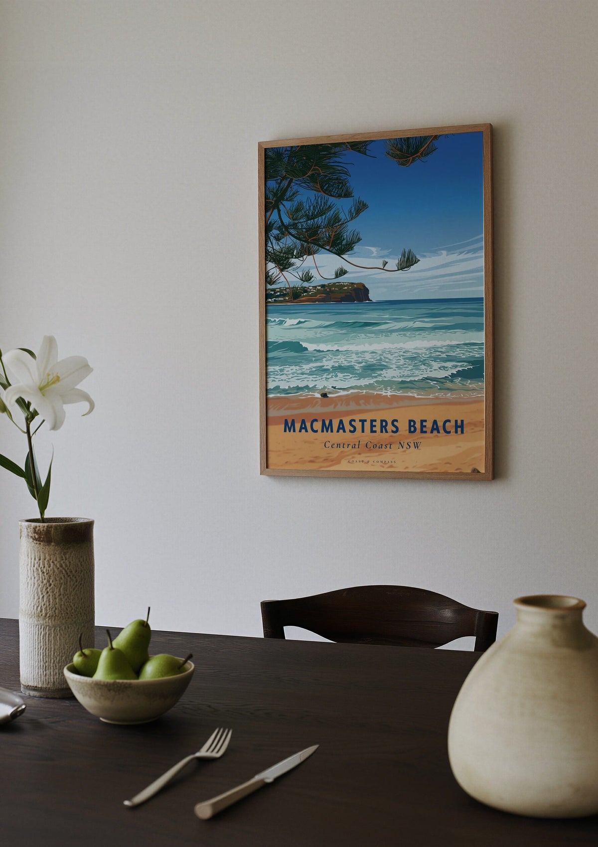 Macmasters Beach travel poster shown above a dining table set with a bowl of pears