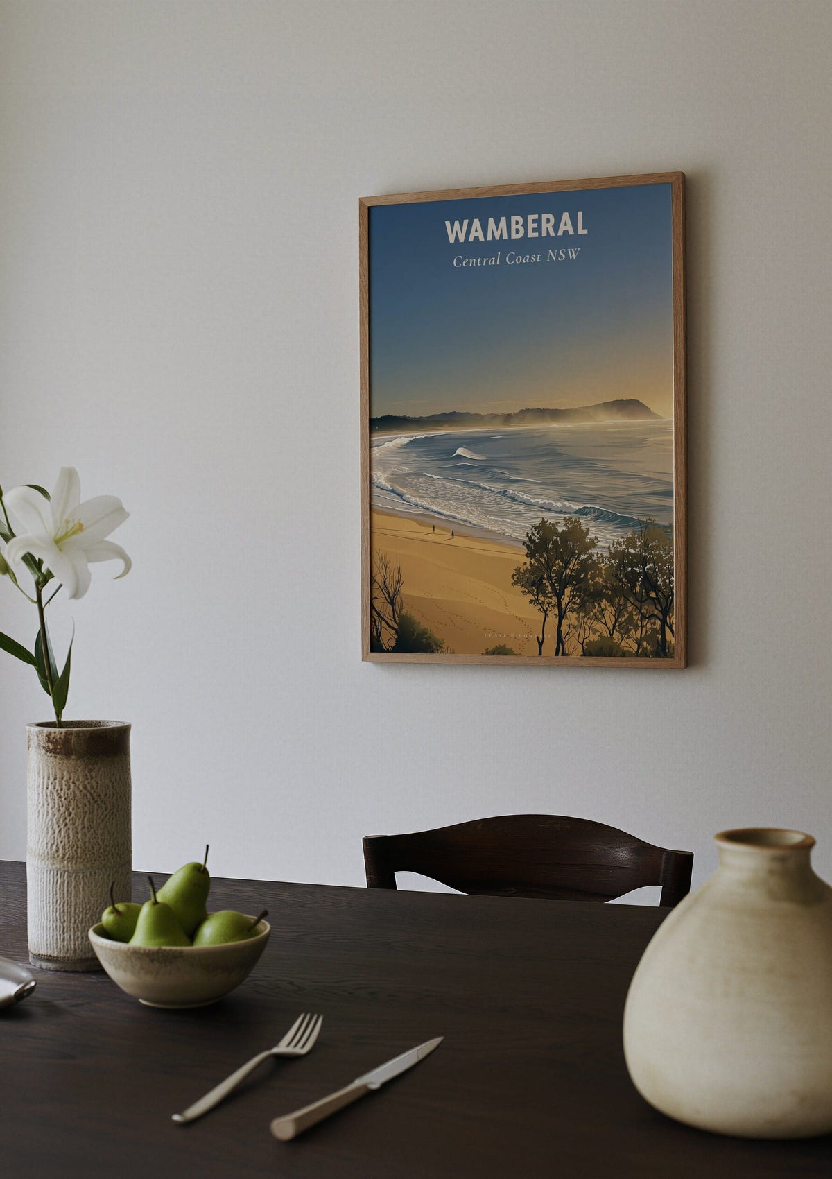 Wamberal Mist travel poster shown above a dining table set with a bowl of pears