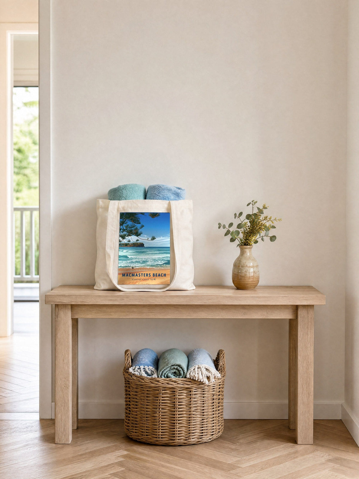 Coast & Compass Macmasters Beach tote bag holding rolled towels on a pale timber console table, beside a ceramic vase of eucalyptus and wattle, with a wicker basket of towels beneath on a herringbone timber floor