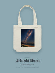 Midnight Bloom artwork printed on a cream cotton canvas tote bag — the Milky Way arcing over a dark coastline