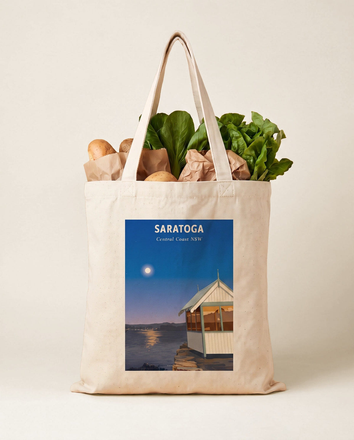 Coast & Compass Saratoga tote bag packed with fresh groceries against a warm sand studio backdrop, cream cotton canvas printed with the Saratoga Central Coast NSW poster design
