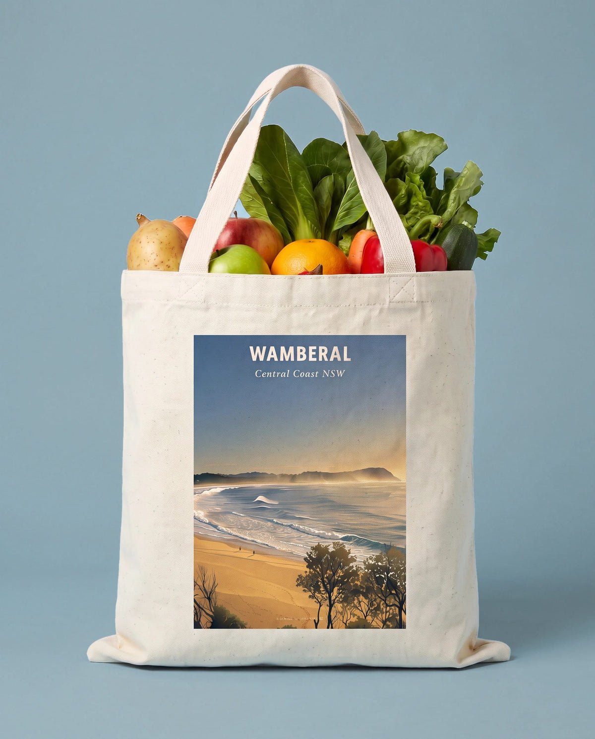 Coast & Compass Wamberal tote bag packed with fresh groceries against a powder blue studio backdrop, cream cotton canvas printed with the Wamberal Central Coast NSW poster design